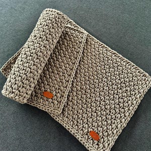 Puede incluir: Una manta y una esterilla de ganchillo color topo hechas a mano. La manta presenta un patrón de puntadas texturizado y está adornada con dos pequeñas etiquetas ovaladas de cuero marrón. Los artículos se muestran sobre una superficie gris oscuro.