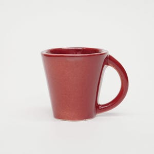 Puede incluir: Una pequeña taza de cerámica de color rojo intenso con un acabado brillante. La taza presenta una forma cónica ligeramente estrecha hacia la base y un asa gruesa y curva unida al lateral. El borde es liso y tiene una apariencia redondeada.