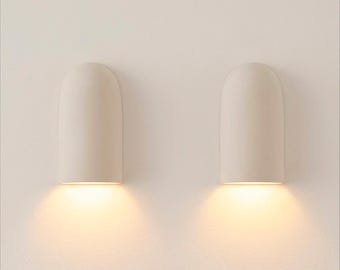 May include: Two off-white, wall-mounted sconces with a rounded top design. Each sconce emits a warm, inviting light downwards, creating a soft glow. The minimalist design is suitable for various interior styles.