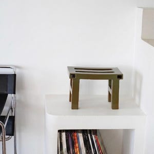 以下が含まれることがあります： 正方形の座面と4本の脚を持つ、オリーブグリーンの小さな木製スツール。スツールは白い曲線の棚の上に置かれています。スツールの下には、雑誌やレコードのコレクションが見えます。左側には黒い椅子があります。スツールの高さは約30cmです。