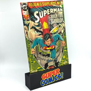 Puede incluir: Un cómic de Superman titulado "Reign of the Supermen!" se muestra en un soporte negro con el texto "SUPER COMICS!". El cómic presenta a Superman con un traje azul y rojo, con una ilustración detallada.