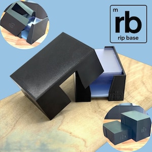 Può includere: Scatola nera per riporre carte con coperchio, contenente una pila di carte. La scatola ha un design unico con il testo "rb rip base" in un quadrato. La scatola è su una superficie di legno.