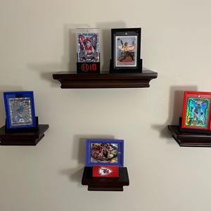 May include: Four different sports cards displayed on three wooden shelves. The shelves are mounted on a white wall. The cards are in protective cases and are displayed vertically. The top shelf has two cards, one in a clear plastic case and the other in a black case. The middle shelf has one card in a blue case. The bottom shelf has one card in a purple case. The cards are all different colors and feature different athletes.