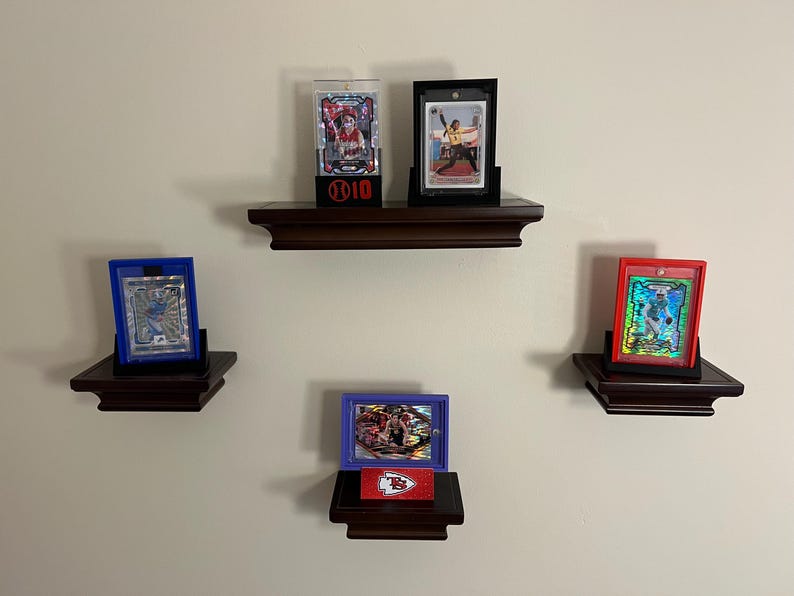 May include: Four different sports cards displayed on three wooden shelves. The shelves are mounted on a white wall. The cards are in protective cases and are displayed vertically. The top shelf has two cards, one in a clear plastic case and the other in a black case. The middle shelf has one card in a blue case. The bottom shelf has one card in a purple case. The cards are all different colors and feature different athletes.