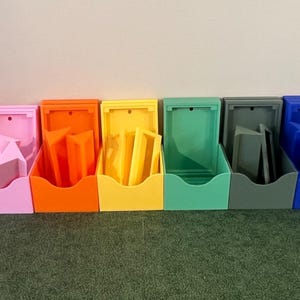 May include: A row of 12 small, open storage boxes in various colors, including white, red, pink, orange, yellow, green, gray, blue, purple, and black. Each box has a small, rectangular opening on the front.