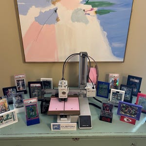 May include: A white and gray 3D printer with a pink platform and a black cable. The printer is surrounded by a variety of framed sports cards, including baseball, football, and basketball cards. The cards are in various colors, including red, blue, and green. The printer is sitting on a light blue table with a white surface. The text "PC CUSTOM" is visible on the front of the printer.