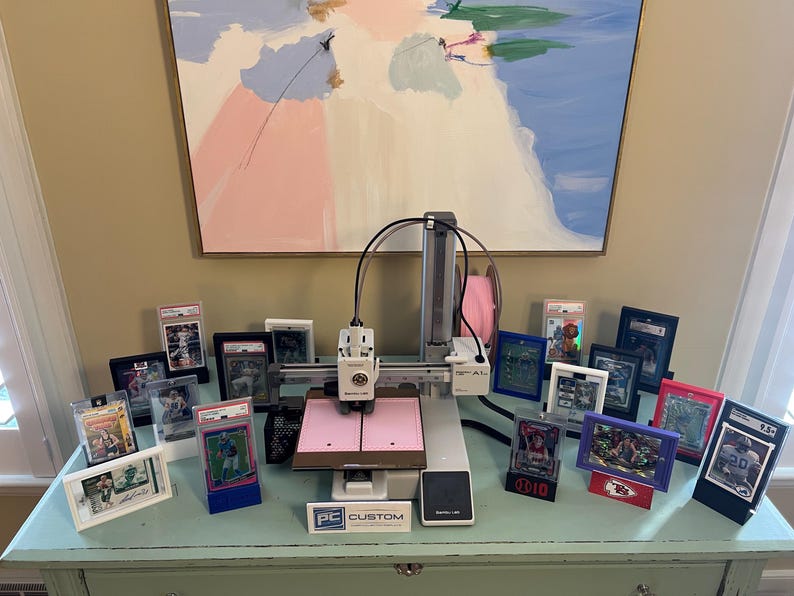 May include: A white and gray 3D printer with a pink platform and a black cable. The printer is surrounded by a variety of framed sports cards, including baseball, football, and basketball cards. The cards are in various colors, including red, blue, and green. The printer is sitting on a light blue table with a white surface. The text "PC CUSTOM" is visible on the front of the printer.
