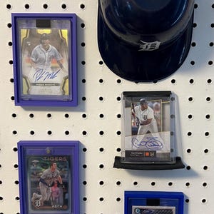 May include: Four baseball cards are displayed on a white pegboard with a blue baseball helmet. The cards are in protective plastic cases with purple and blue borders. The cards feature baseball players from the Detroit Tigers. The cards feature the names "Pasquel Meadows", "Casey Fielder", "Keith", and "Jake Rogers".