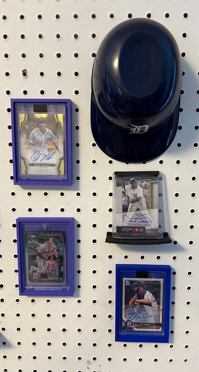 May include: Four baseball cards are displayed on a white pegboard with a blue baseball helmet. The cards are in protective plastic cases with purple and blue borders. The cards feature baseball players from the Detroit Tigers. The cards feature the names "Pasquel Meadows", "Casey Fielder", "Keith", and "Jake Rogers".