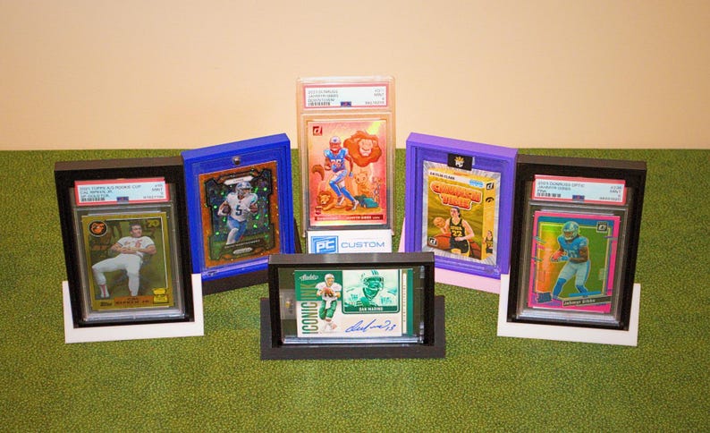 May include: Six different sports trading cards are displayed in protective cases. The cases are black, blue, purple, and clear. The cards feature athletes in various sports, including football and basketball. The cards are graded by PC Custom.