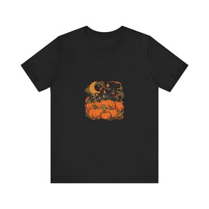 Puede incluir: Una camiseta negra de manga corta con una ilustración gráfica central de un gato negro descansando sobre un grupo de calabazas naranjas. El diseño incluye una luna creciente amarilla, estrellas pequeñas y follaje verde oscuro.