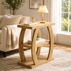 May include: A light oak wood side table featuring a rectangular top, a middle shelf, and a solid base. The table is supported by two curved, C-shaped side panels. A small lamp, a glass of water, and a reed diffuser sit on the top surface.