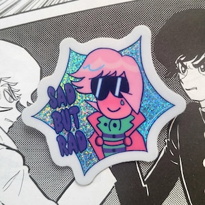 以下が含まれることがあります： ピンクの髪とサングラスをした漫画のキャラクターが特徴のホログラフィックステッカー。キャラクターは緑と紫の服とピンクのマントを着ています。ステッカーには紫色のテキストで「SAD BUT RAD」と書かれています。