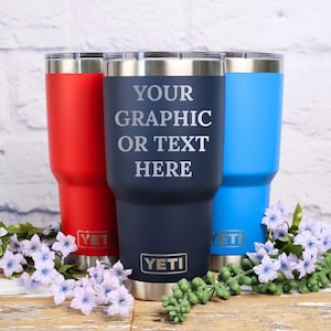 May include: Three insulated tumblers in red, navy blue, and blue. The navy blue tumbler has the text "YOUR GRAPHIC OR TEXT HERE". Each tumbler has a silver rim and the "YETI" logo. The tumblers are decorated with purple flowers.