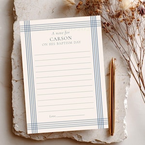 May include: A note card with the text "A note for Carson on his Baptism Day" in a serif font. The card has a cream background with blue striped borders and lined paper. A gold pen and dried flowers are next to the card.