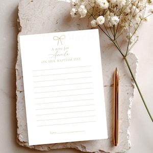 May include: A white rectangular stationery card featuring a gold line drawing of a bow at the top, followed by the text A note for Amelia ON HER BAPTISM DAY, with horizontal lines for writing and a Love line at the bottom, accompanied by a gold pen.