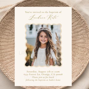May include: A baptism invitation card featuring a photo of a young girl with long hair, framed by a decorative border. The card includes the text "You're invited to the baptism of Landrie Kate" and event details in a soft, elegant design. The card is placed on a woven plate.