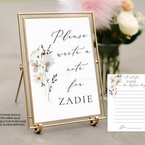 May include: A white sign with a gold frame and easel, featuring a floral design and the text "Please write a note for Zadie". A matching white card with a floral design and lines for writing a note is also included.