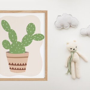 May include: A framed print of a green cactus in a brown pot with a geometric pattern. The print is on a light pink background and is surrounded by a light wood frame.