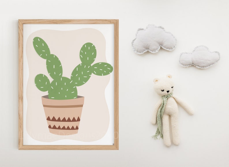 May include: A framed print of a green cactus in a brown pot with a geometric pattern. The print is on a light pink background and is surrounded by a light wood frame.