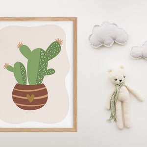 May include: A framed print of a green cactus in a brown pot with a heart on the front. The print is on a white background with two white clouds and a white crocheted bear on the right side of the image.