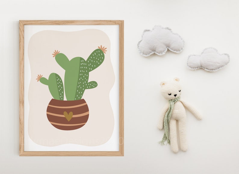May include: A framed print of a green cactus in a brown pot with a heart on the front. The print is on a white background with two white clouds and a white crocheted bear on the right side of the image.