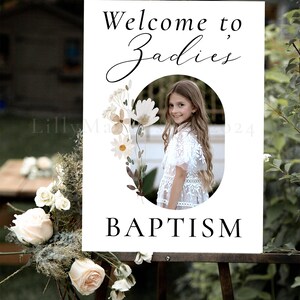 May include: White welcome sign with a floral design and a photo of a girl. The sign says "Welcome to Zadies Baptism".