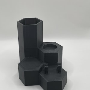 May include: A black, multi-level organizer with a hexagonal design. The organizer has a tall, open compartment, a smaller circular area, and a section with two small posts. The item is made of a textured material.