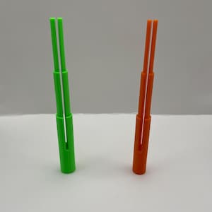 Puede incluir: Dos herramientas de plástico, una verde y otra naranja, se muestran sobre un fondo neutro. Cada herramienta tiene una base cilíndrica con dos varillas verticales, delgadas y paralelas, que se extienden hacia arriba. Las varillas están separadas por un pequeño espacio.