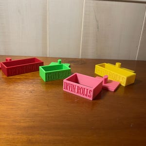 May include: Four small, rectangular containers in red, green, pink, and yellow, each with the text "IRVIN ROLLS" embossed on the side. The containers are arranged on a wooden surface, with a light-coloured wall in the background.