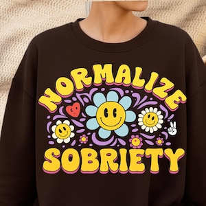 Puede incluir: Una sudadera marrón oscuro con las palabras "NORMALIZE SOBRIETY" en amarillo, con un diseño de flores y caritas sonrientes. El diseño incluye una flor azul con una carita sonriente, corazones y otras flores. La sudadera es de estilo casual.