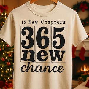 Pode incluir: Uma camiseta creme com o texto "12 New Chapters, 365 new chance" em preto. A camiseta está pendurada em um cabide de madeira. Uma árvore de Natal e uma lareira são visíveis ao fundo.
