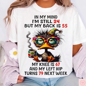 Puede incluir: Una camiseta blanca con el gráfico de un pato despeinado que lleva un sombrero de fiesta colorido y gafas grandes, sosteniendo una taza humeante. El texto dice: IN MY MIND I'M STILL 24 BUT MY BACK IS 55 MY KNEE IS 67 AND MY LEFT HIP TURNS 79 NEXT WEEK.