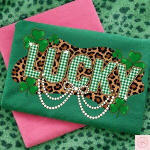 May include: Green t-shirt with the word "LUCKY" in a green and white checkered pattern, set against a leopard print background. The design includes shamrocks and a pearl necklace. A pink shirt is folded underneath.