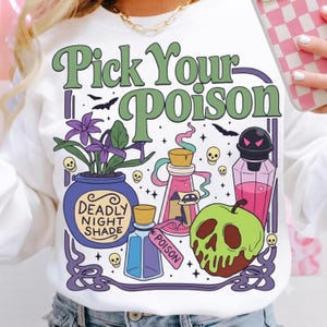 May include: A white sweatshirt featuring a graphic print with the text Pick Your Poison. The design includes a purple pot labeled Deadly Night Shade, a pink potion bottle, a blue bottle labeled Poison, a green skull-shaped apple, and bats.