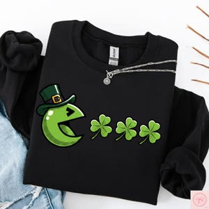 Könnte beinhalten: Schwarzes Sweatshirt mit einem grünen Leprechaun-ähnlichen Grafik und drei Kleeblättern. Eine silberne Halskette mit Anhänger liegt über dem Sweatshirt. Das Sweatshirt befindet sich auf einer Jeansoberfläche.