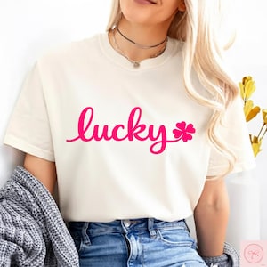 Könnte beinhalten: Cremefarbenes T-Shirt mit dem Wort "lucky" in leuchtend pinkfarbener Schrift, begleitet von einer pinkfarbenen vierblättrigen Kleeblatt-Grafik. Das Shirt hat einen Rundhalsausschnitt und kurze Ärmel. Ein grauer Pullover ist über den Schultern drapiert.