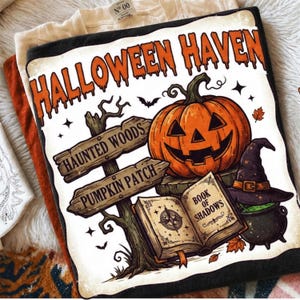 May include: A black t-shirt featuring a graphic print with the text HALLOWEEN HAVEN, HAUNTED WOODS, PUMPKIN PATCH, and BOOK OF SHADOWS. The design includes a carved orange pumpkin, a wooden signpost, an open book, and a witch hat on a cauldron.