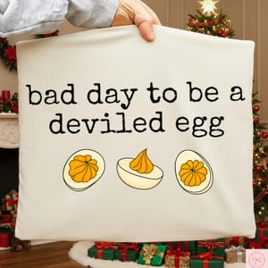 Può includere: Cuscino color crema con la scritta nera "bad day to be a deviled egg". Sotto, tre illustrazioni di uova alla diavola con ripieno giallo. Il cuscino è mostrato davanti a un albero di Natale.