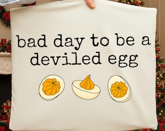 May include: Cream-colored pillow with the phrase "bad day to be a deviled egg" in black text. Below the text are three illustrations of deviled eggs with yellow filling. The pillow is held up in front of a Christmas tree.