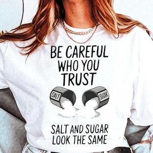 以下が含まれることがあります： 「BE CAREFUL WHO YOU TRUST」の文字がプリントされた白いTシャツ。デザインには、「SALT」と「SUGAR」とラベルされた2つのひっくり返った容器と、こぼれた内容物が含まれています。「SALT AND SUGAR LOOK THE SAME」というフレーズが下に印刷されています。