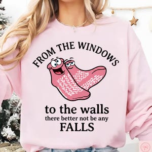 以下が含まれることがあります： 「FROM THE WINDOWS to the walls there better not be any FALLS」の文字が入った薄いピンク色のスウェットシャツ。ピンク色の顔と白い模様の靴下2足のグラフィックが特徴です。