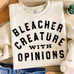 May include: Cream-colored sweatshirt with the text "BLEACHER CREATURE WITH OPINIONS" in bold, black letters. The sweatshirt is folded, with a glimpse of denim jeans at the bottom. A hat and a small plant are visible in the background.