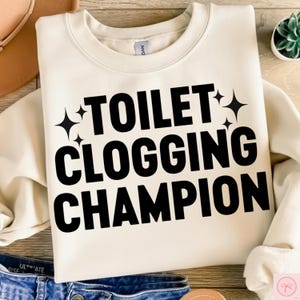 May include: Cream-colored sweatshirt with the text "TOILET CLOGGING CHAMPION" in bold black letters. The text is accented with four black starburst graphics. The sweatshirt is folded and displayed on a light-colored surface.