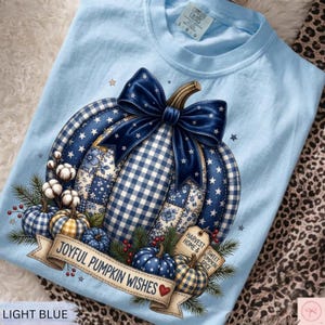 Könnte beinhalten: Ein hellblaues T-Shirt mit einer großen Grafik eines Kürbisses, der mit blau-weißem Vichy-Muster, Blumen- und Sternenmustern verziert ist. Eine dunkelblaue Schleife sitzt auf dem Kürbis, der von Baumwollblüten, Tannennadeln und Beeren umgeben ist. Ein Banner trägt die Aufschrift JOYFUL PUMPKIN WISHES, mit Schildern HARVEST HOME und SWEET PUMPKIN SPICE.