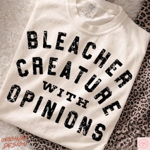 Puede incluir: Camiseta color crema con la frase "BLEACHER CREATURE WITH OPINIONS" en letras negras desgastadas. La camiseta tiene cuello redondo y mangas cortas. Las palabras "ORIGINAL DESIGN" están impresas en la parte inferior izquierda.