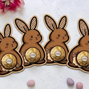 Könnte beinhalten: Vier braune, hasenförmige Ausstecher mit gemusterten Ohren und einem goldverpackten Schokoladenbonbon in der Mitte. Die Hasen haben einen dekorativen, gewellten Rand und sind auf einer weißen Oberfläche mit Osterdekorationen angeordnet.