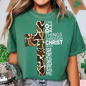 May include: A green t-shirt featuring a cross design with a camouflage pattern in shades of brown and green. The text on the shirt reads "I CAN DO ALL THINGS THROUGH CHRIST WHO STRENGTHENS ME" in white lettering.