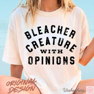 May include: Cream-colored t-shirt with the text "BLEACHER CREATURE WITH OPINIONS" in a bold, black font. The words "ORIGINAL DESIGN" are printed in a rust-colored font at the bottom left.
