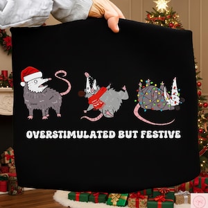 May include: Black fabric featuring three cartoon opossums in festive poses. One wears a Santa hat, another a scarf, and the last is tangled in Christmas lights. The text "OVERSTIMULATED BUT FESTIVE" is displayed in white.